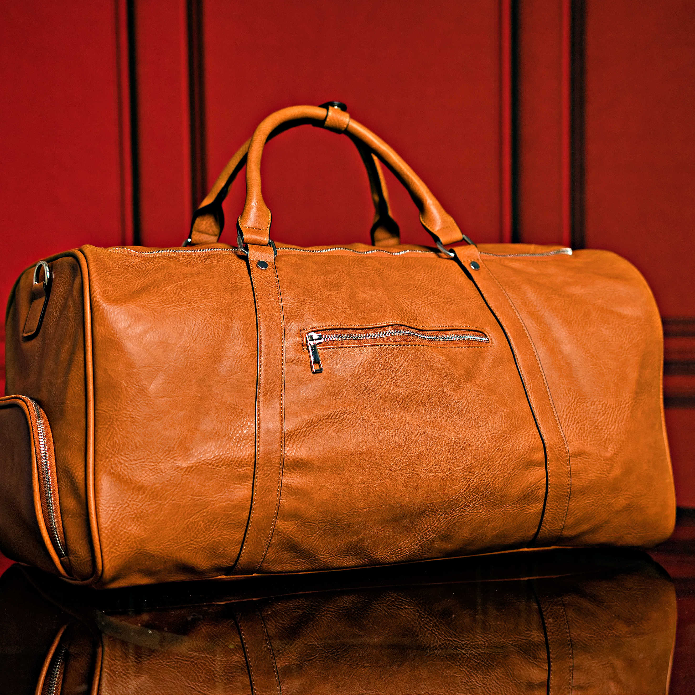 Brown leather duffel bag on a reflective surface with a red curtain background