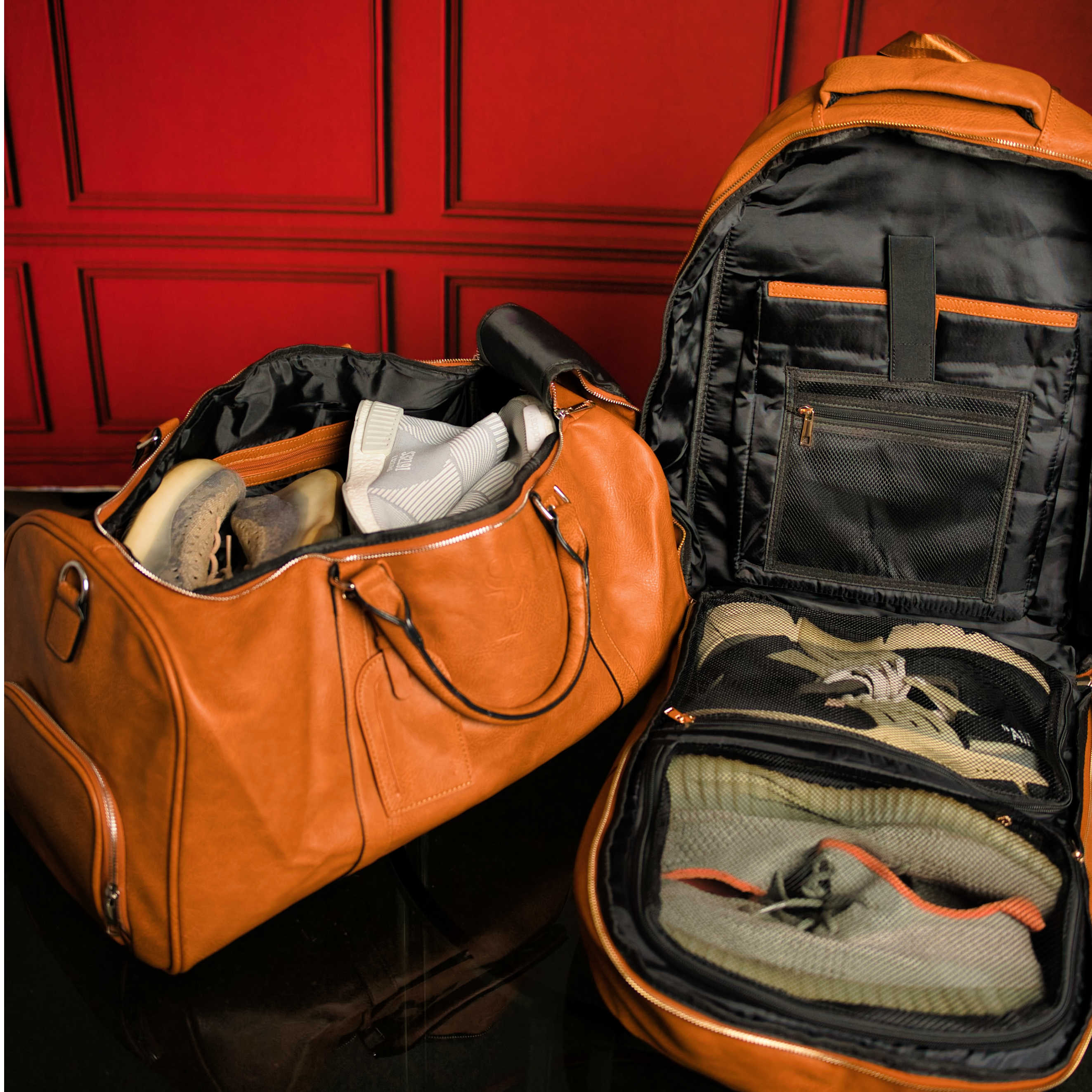 Orange duffel bag and black backpack with shoes and clothes inside against a red paneled wall.