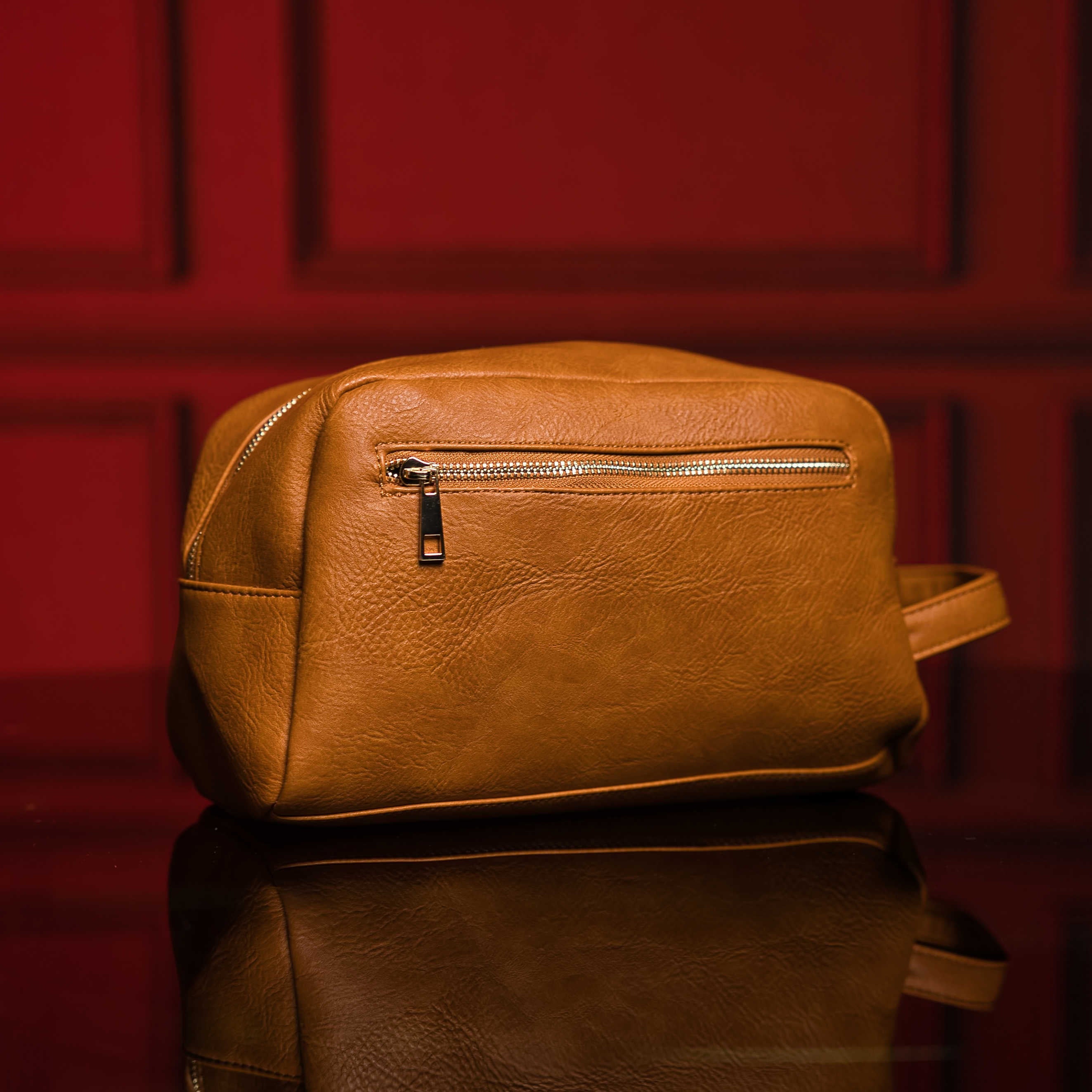 Brown leather toiletry bag with a zipper on a reflective surface with a red background