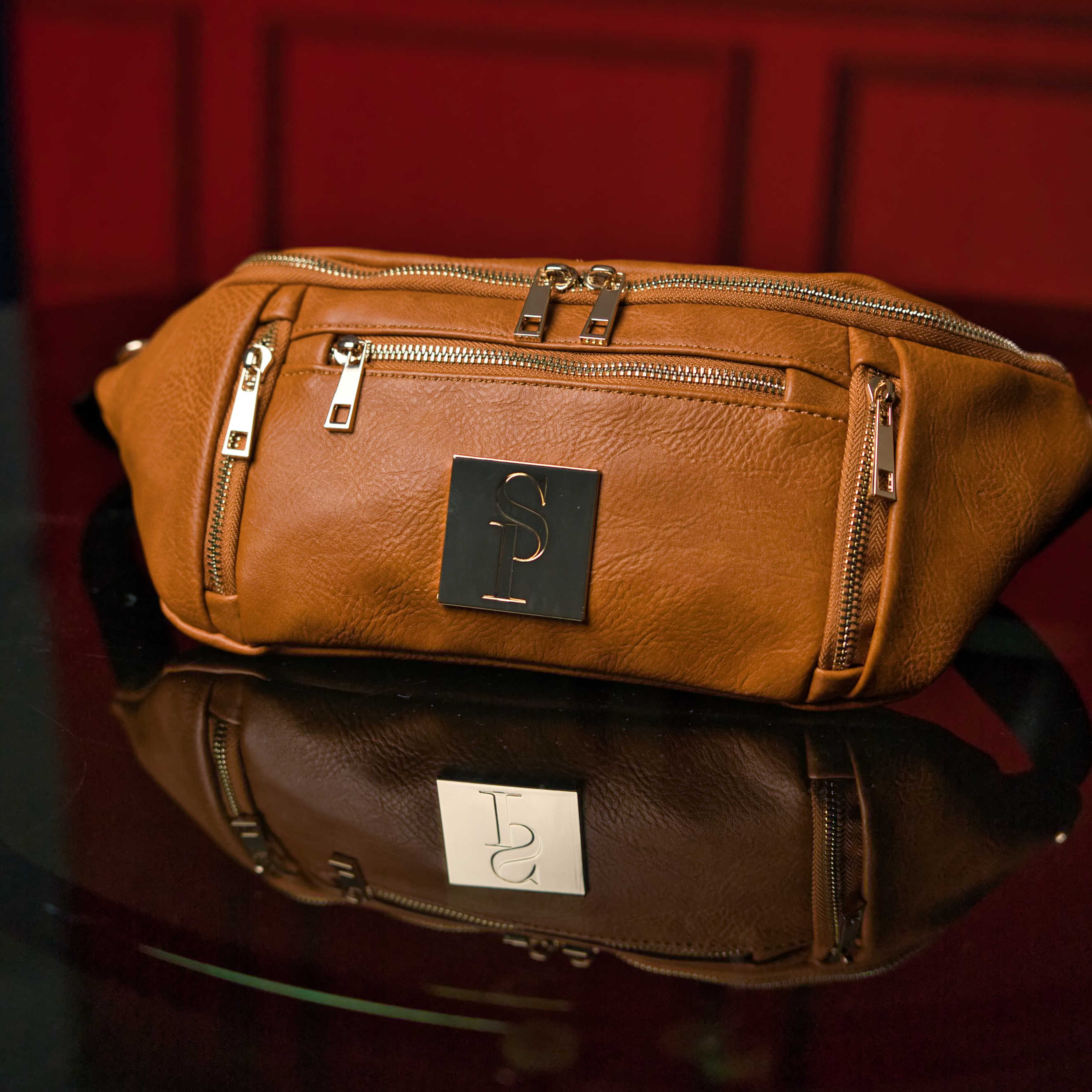 Brown leather waist bag with gold zippers on a reflective surface with a red background