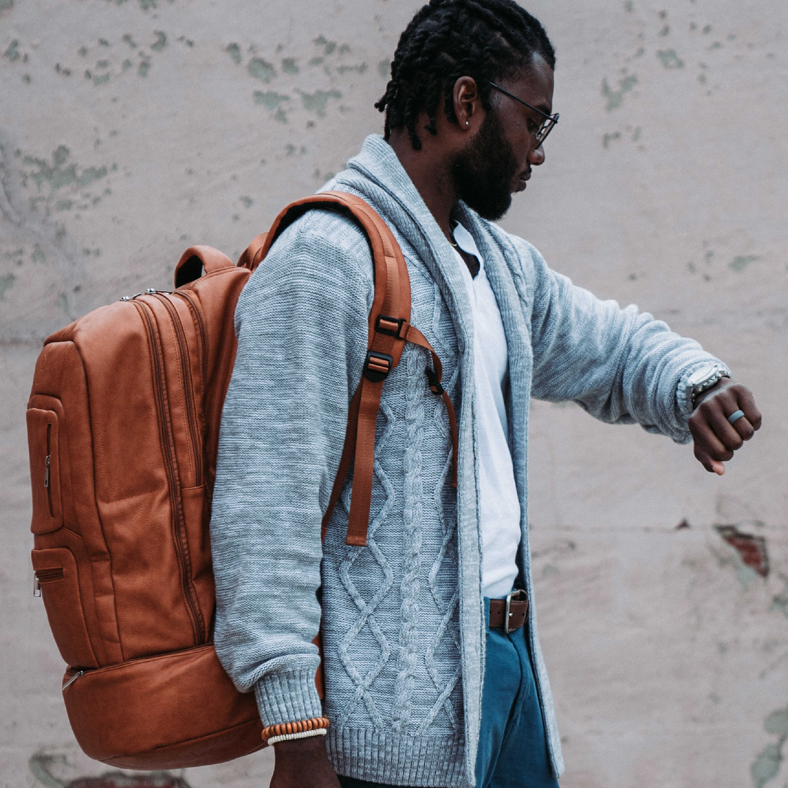 Man wearing a brown leather backpack against a textured wall.