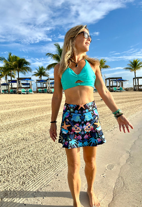 Woman in turquoise top and colorful skirt standing on a beach with palm trees and boats in the background.