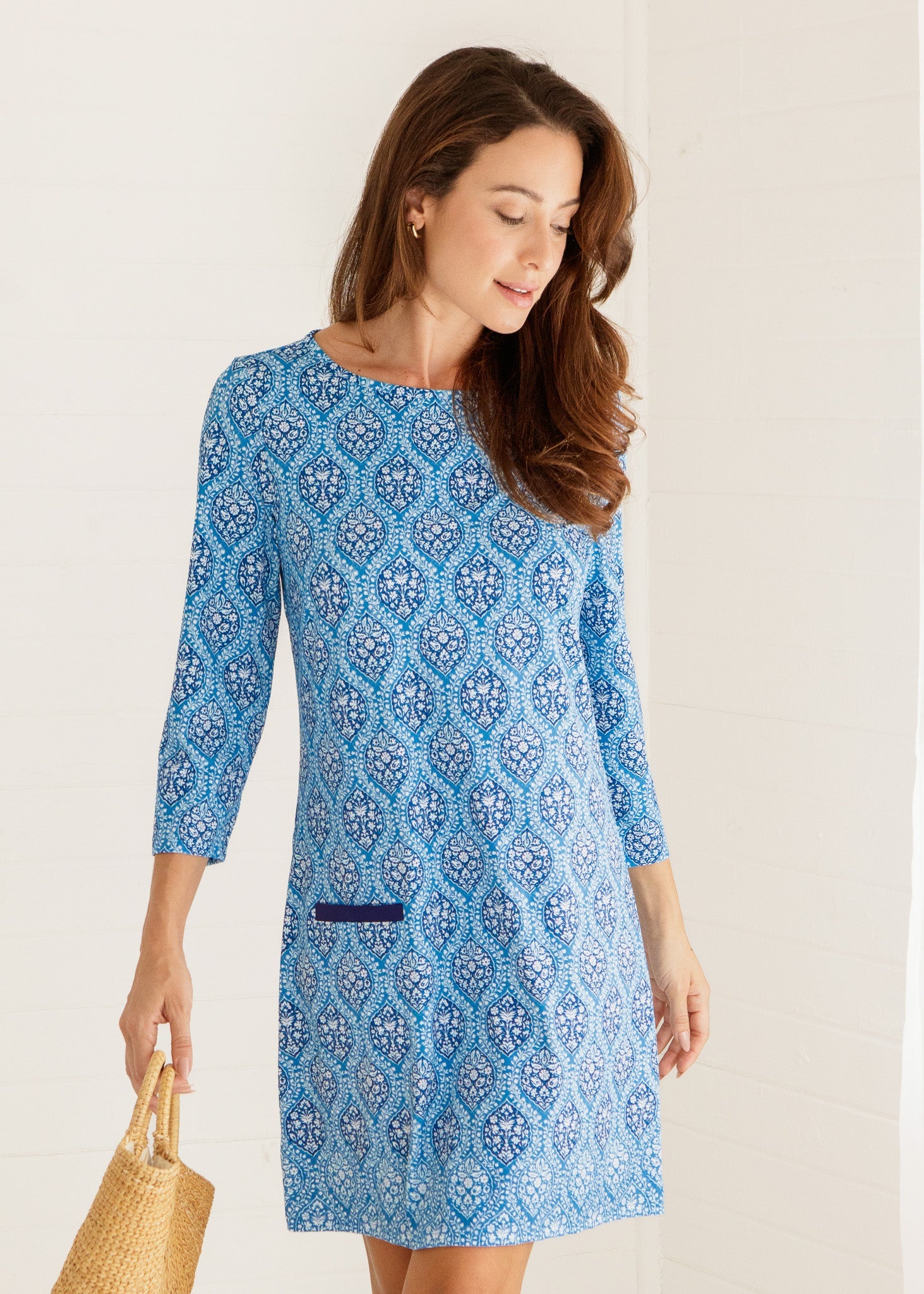 Woman wearing a blue patterned dress holding a straw bag against a plain background