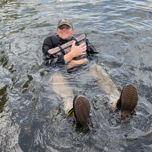 Person sitting in water holding a large fish, wearing boots and a cap.