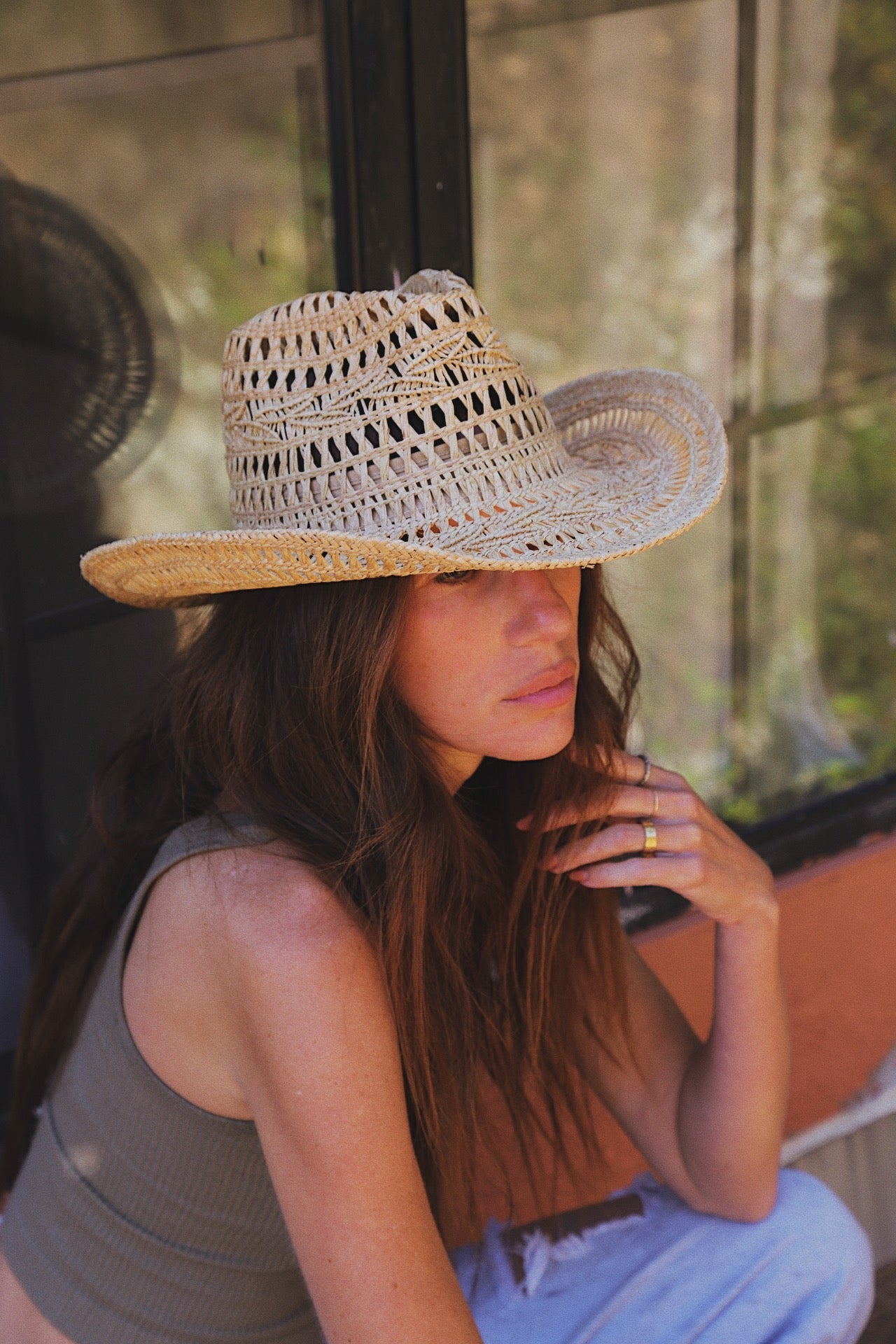 Woman wearing a straw hat sitting by a window with a blurred outdoor background