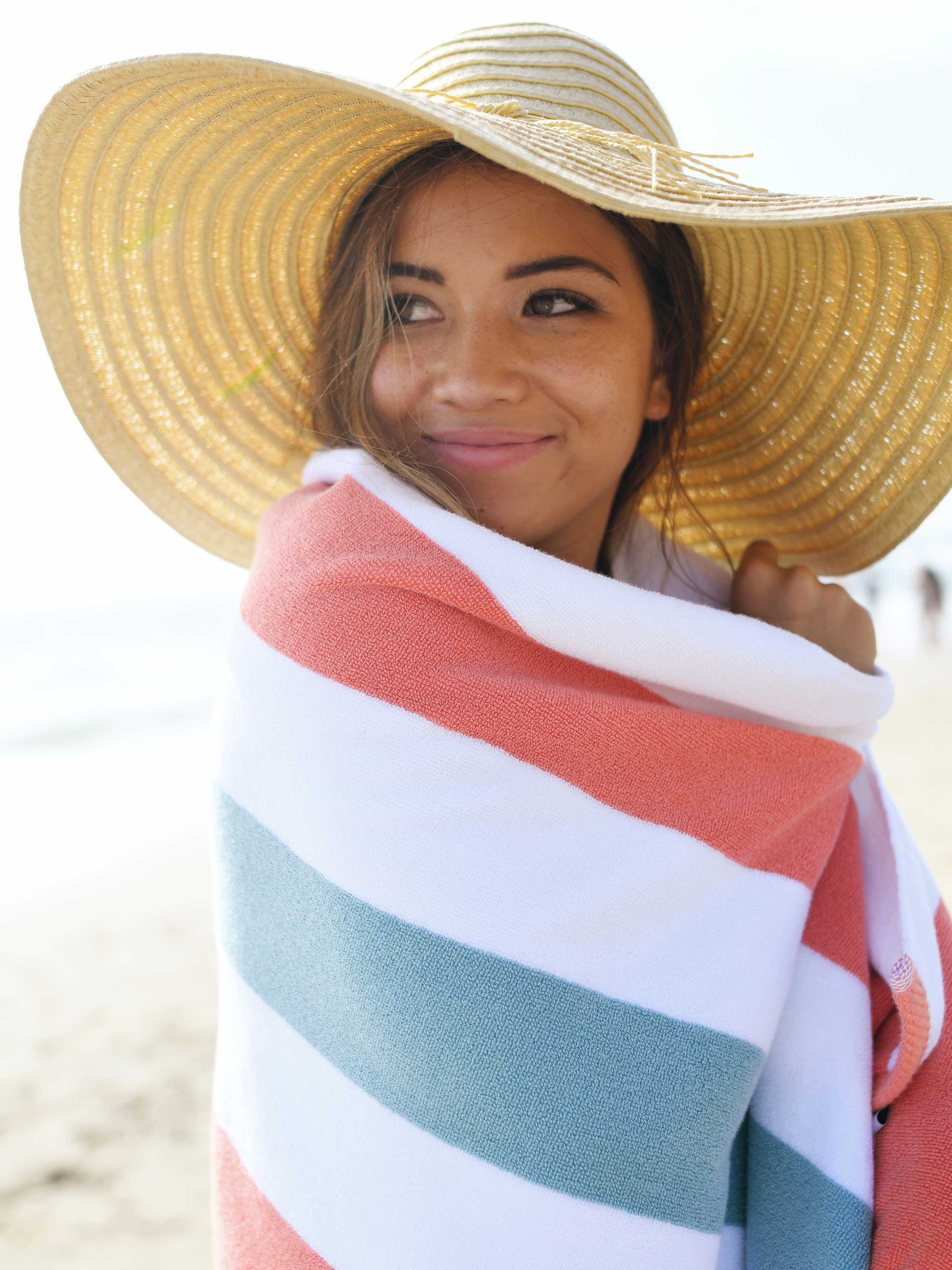Woman wrapped in a striped towel wearing a wide-brimmed hat on a beach.