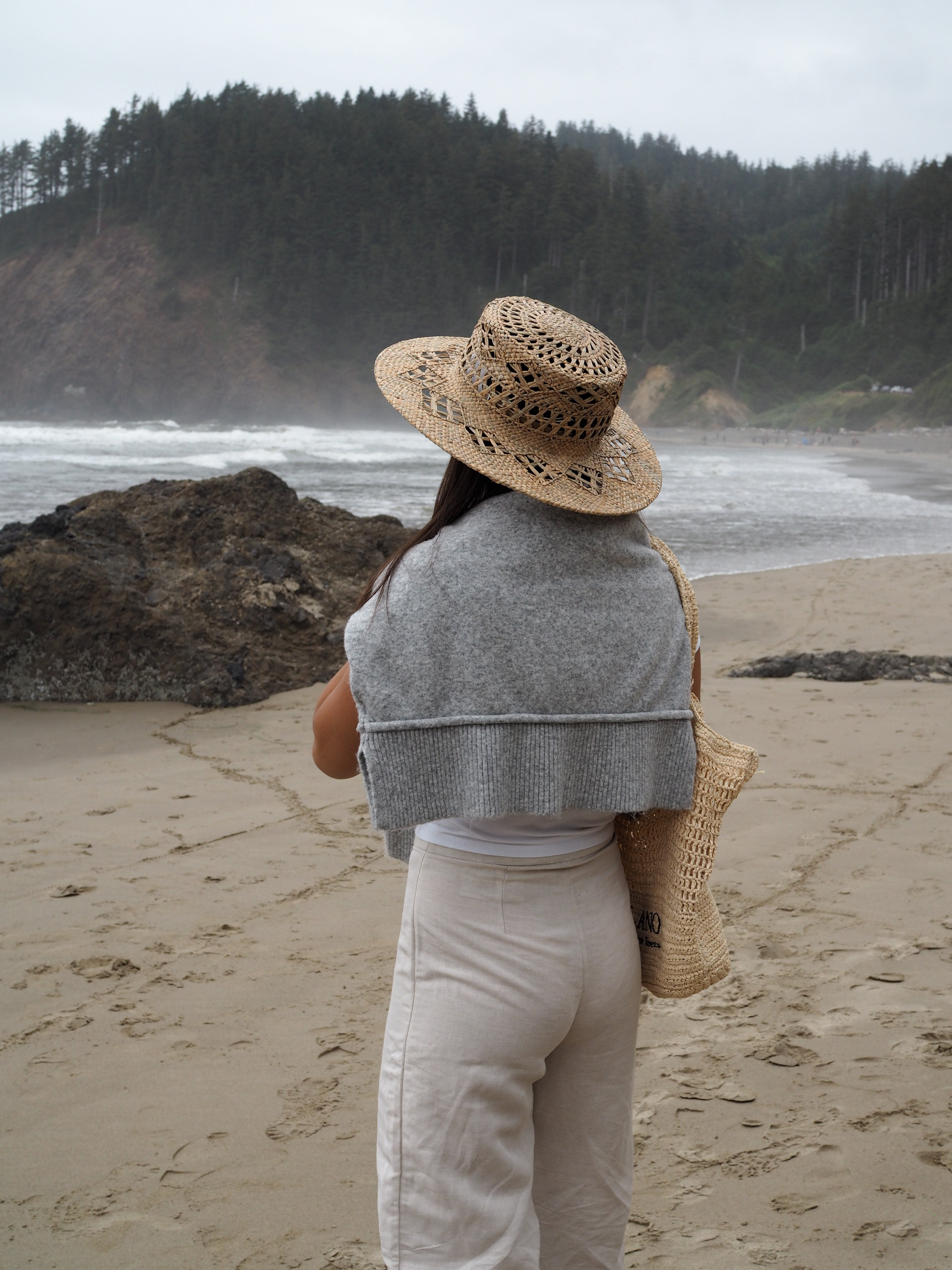 Person standing on a beach holding a hat, with a forested hill in the background.