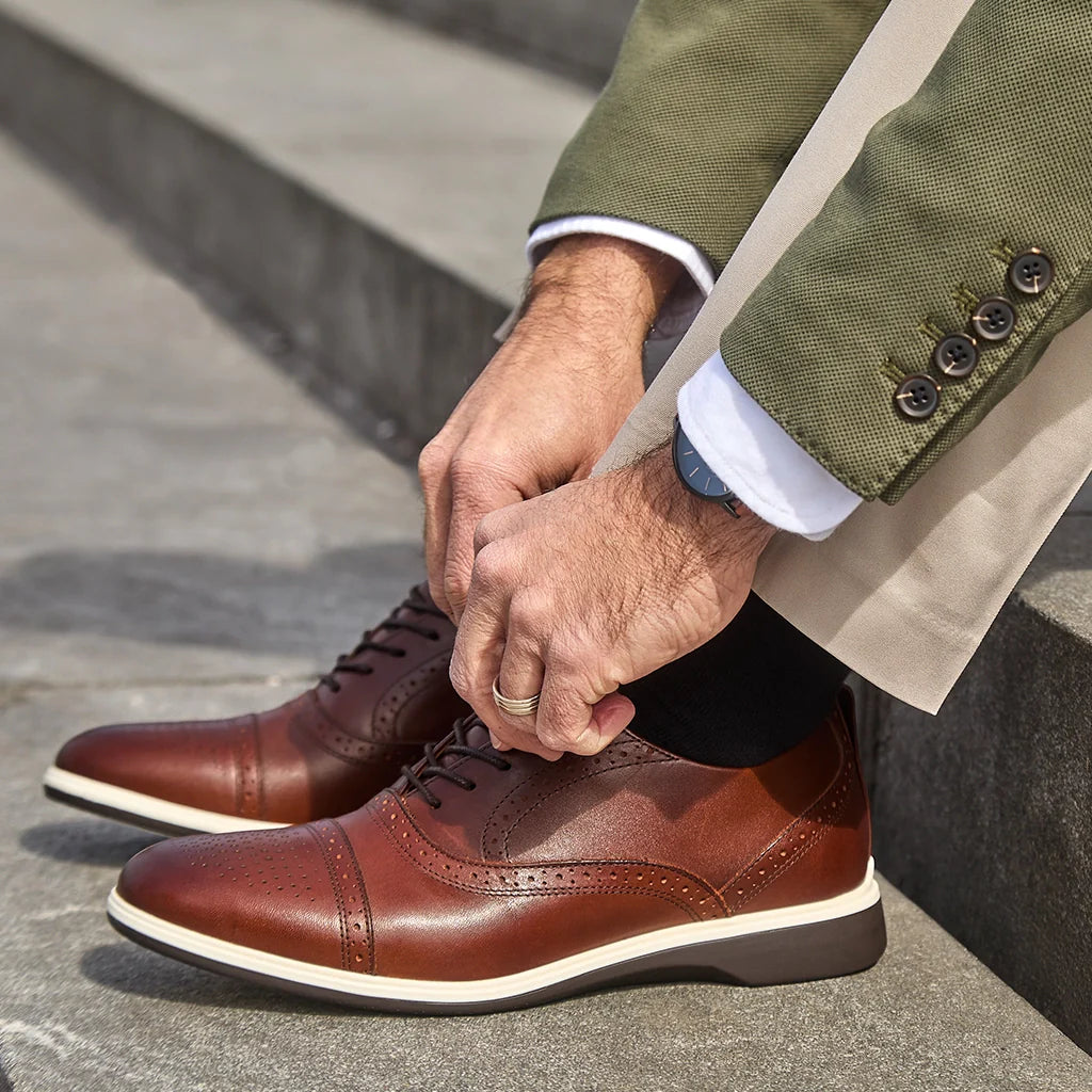 Person wearing brown leather shoes and a green suit adjusting their shoe outdoors.
