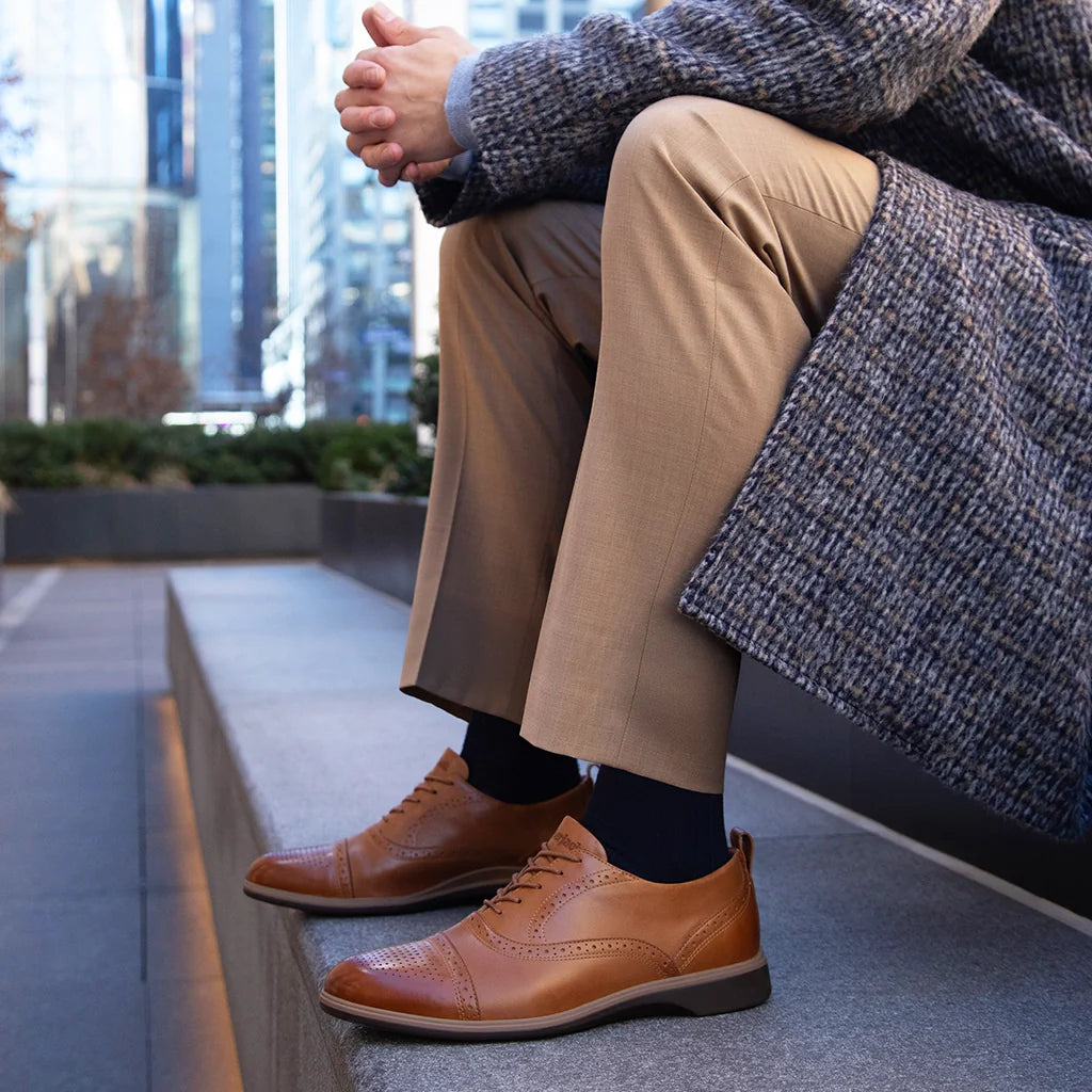 Person wearing brown shoes and beige pants sitting on a ledge with a cityscape background