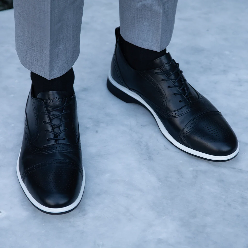 Black leather shoes with white soles worn by a person on a light gray background