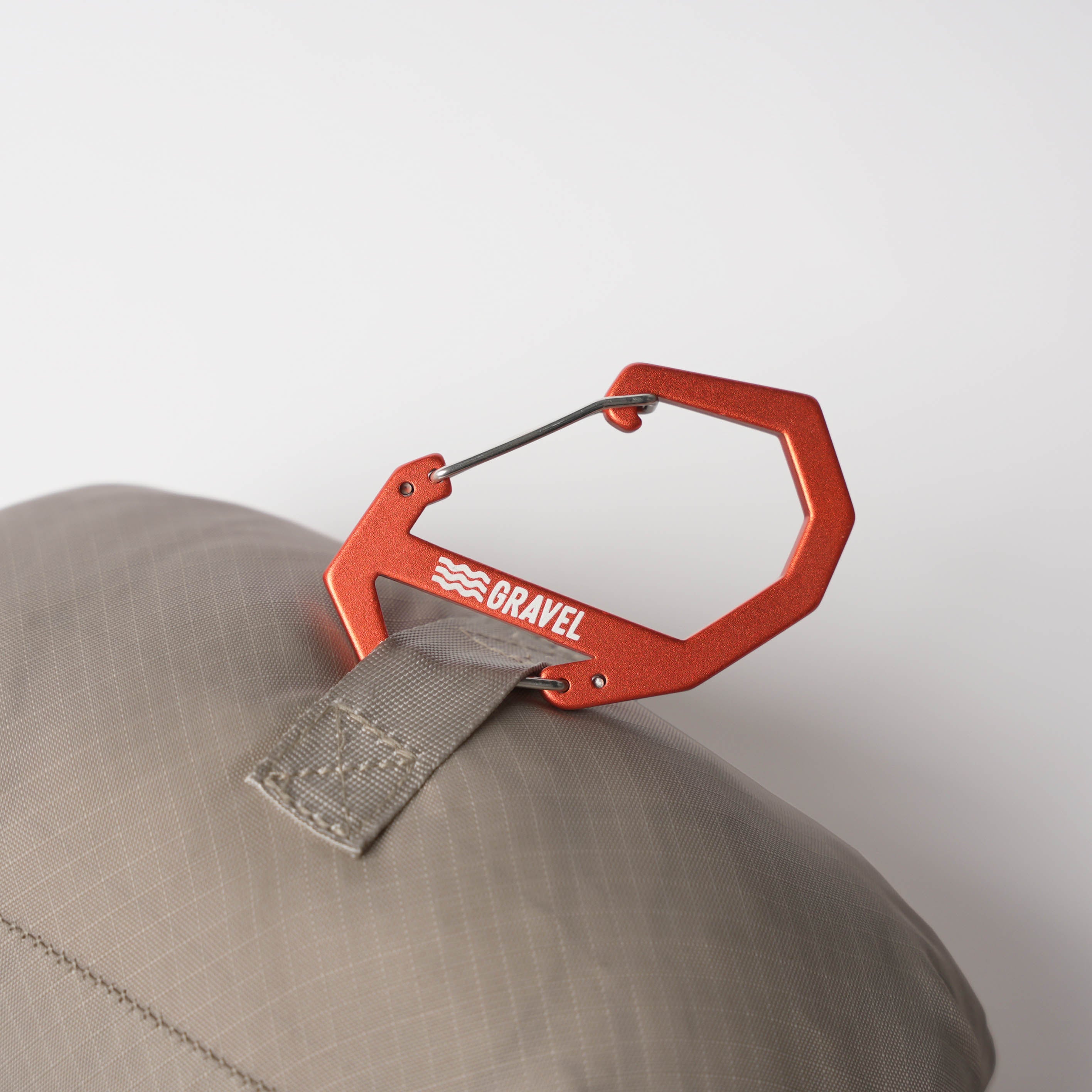 Red carabiner with 'Gravel' branding attached to a beige cap on a white background