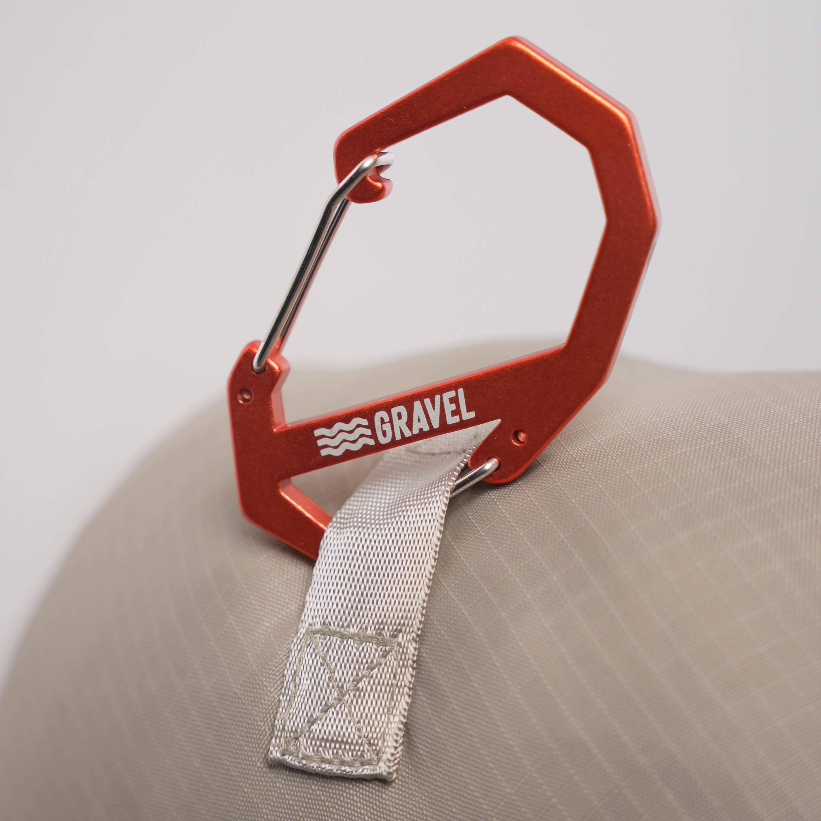 Red carabiner with a strap attached, branded 'Gravel', on a beige surface.
