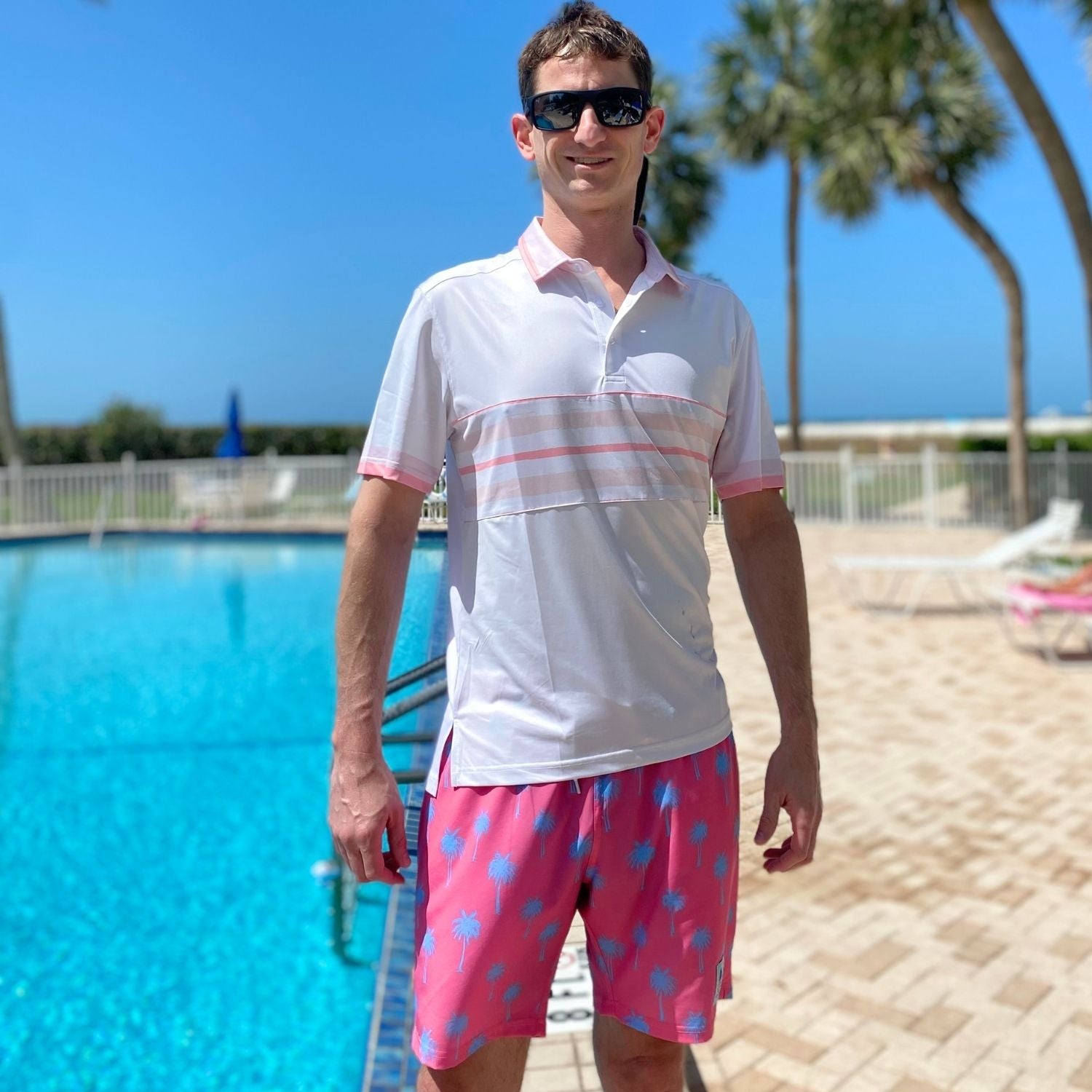 Man wearing a white polo shirt with pink accents and pink shorts with blue patterns by a pool.