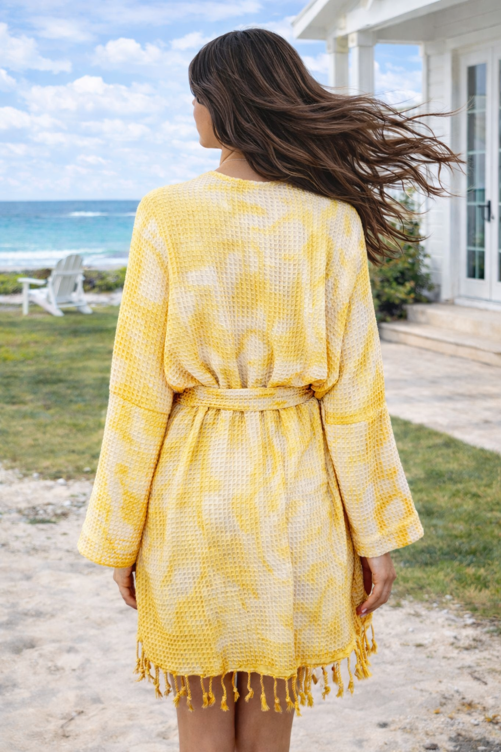 Woman in a yellow patterned dress standing on a beach with ocean and house in the background