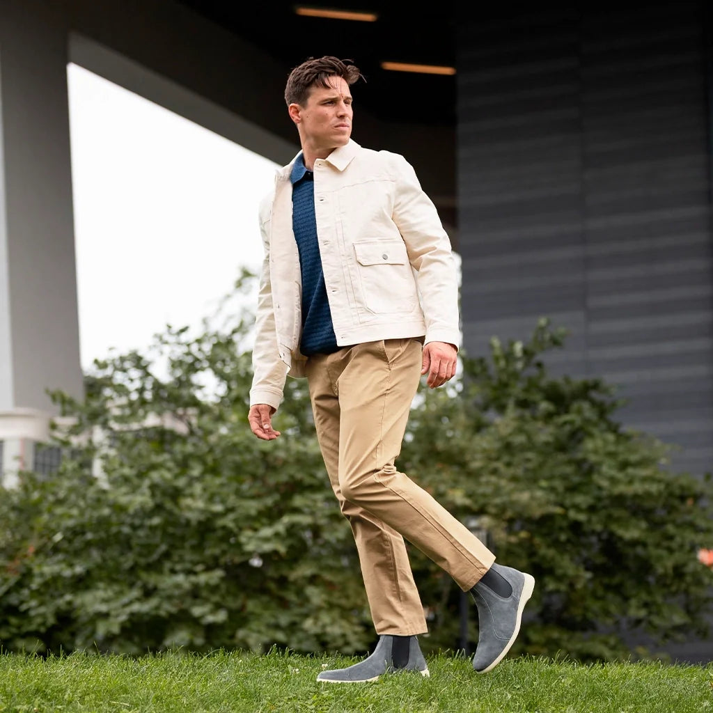 Man walking outdoors wearing a cream jacket, navy shirt, beige pants, and gray shoes.