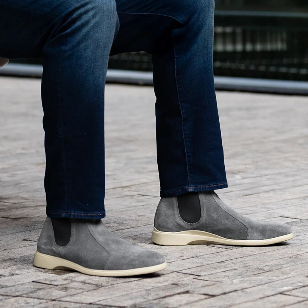 Person wearing gray suede Chelsea boots with dark blue jeans on a stone pavement.