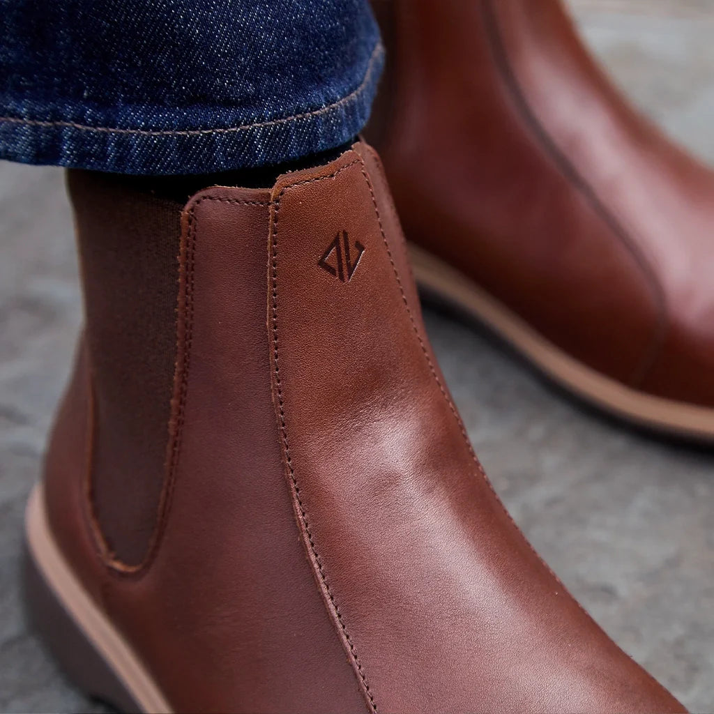 Brown leather boots with a brand logo worn with blue jeans on a gray surface
