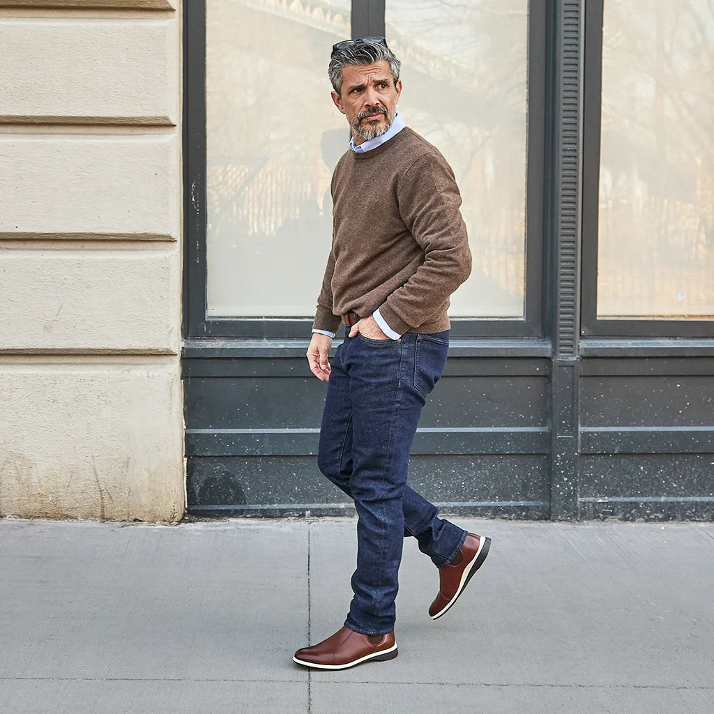 Man wearing a brown sweater and dark jeans standing against a building.