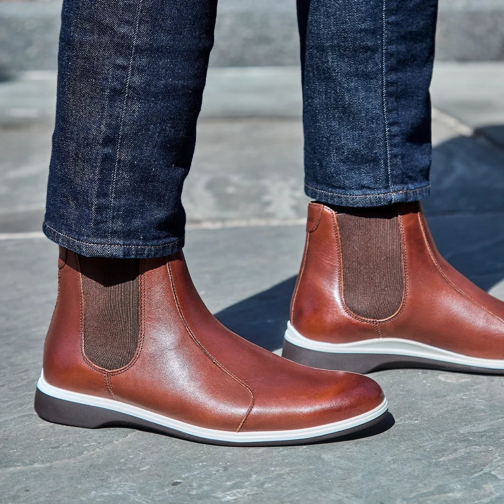 Brown leather boots with white soles worn with blue jeans on a gray pavement.