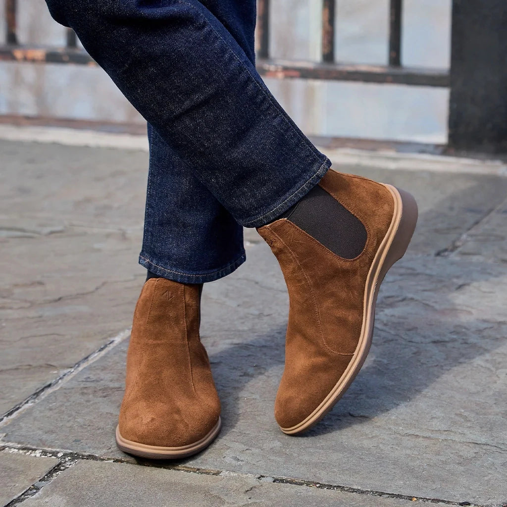 Brown suede shoes worn with rolled-up jeans on a stone pavement.