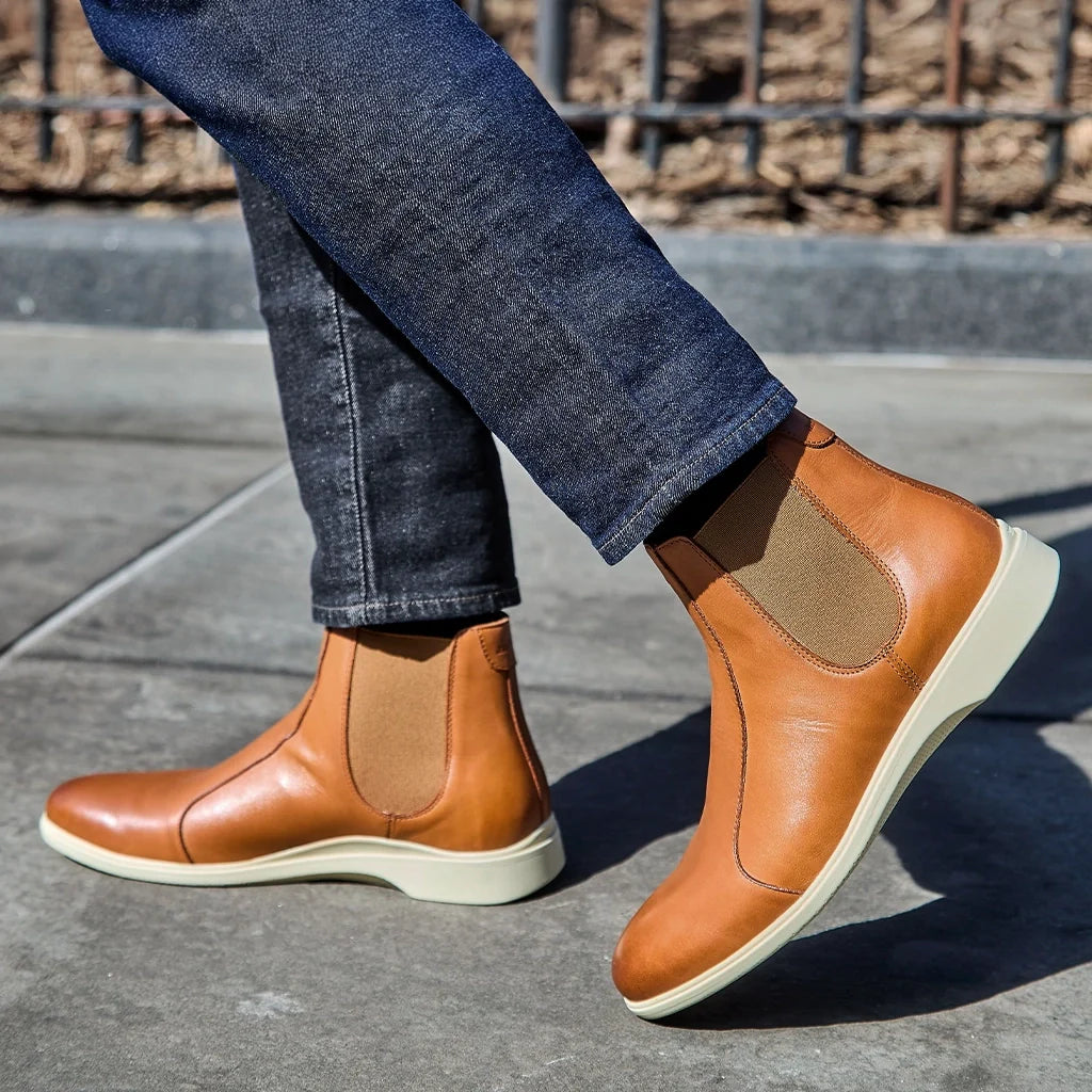 Brown Chelsea boots worn with blue jeans on a concrete surface.