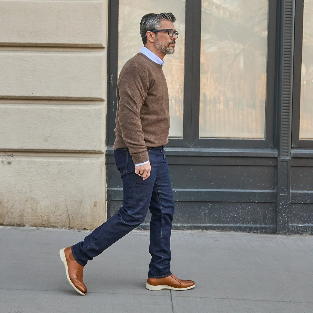 Man walking on a sidewalk wearing a brown sweater, blue jeans, and brown shoes.