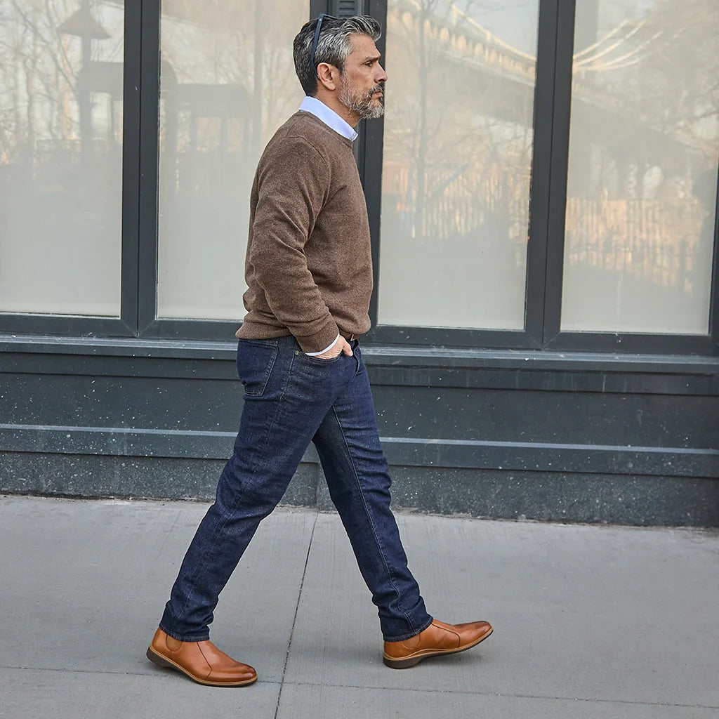 Man walking on a sidewalk wearing a brown sweater, blue jeans, and brown shoes.