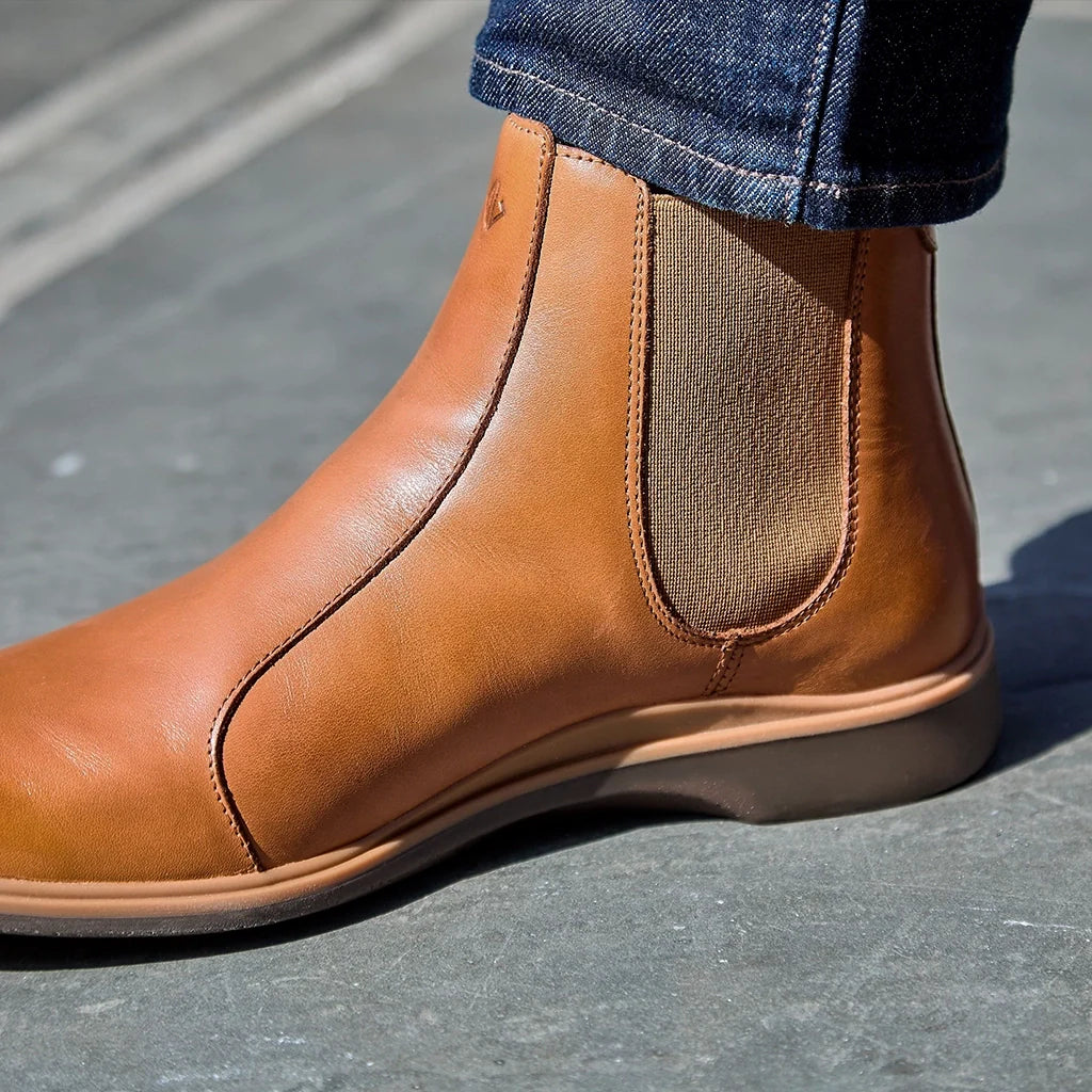 Brown leather Chelsea boot worn with blue jeans on a gray pavement.