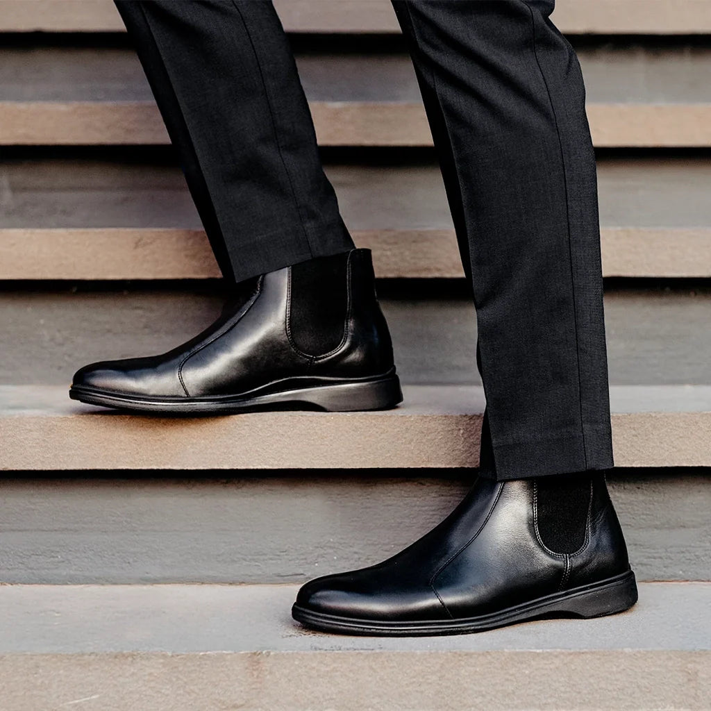 Person wearing black leather Chelsea boots and dark pants on wooden steps.