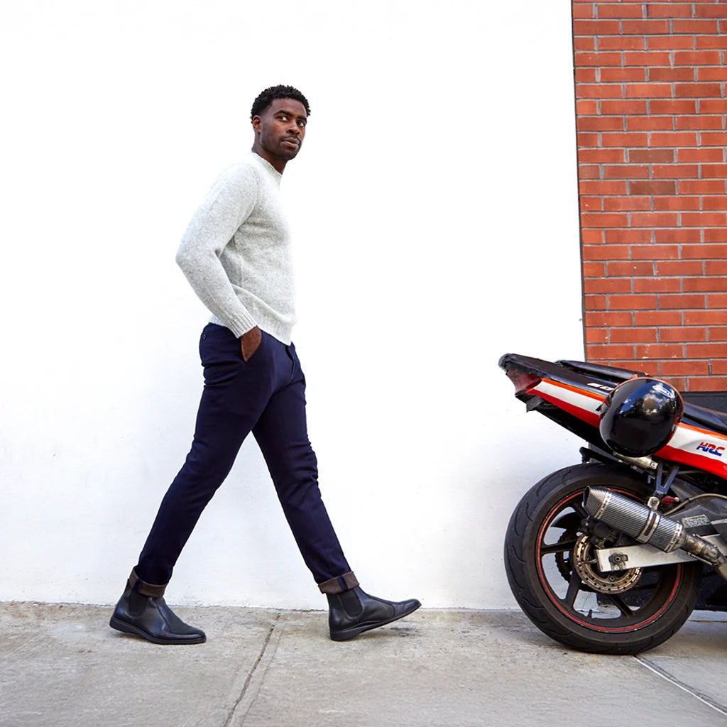 Man walking next to a motorcycle against a white wall with a brick corner.
