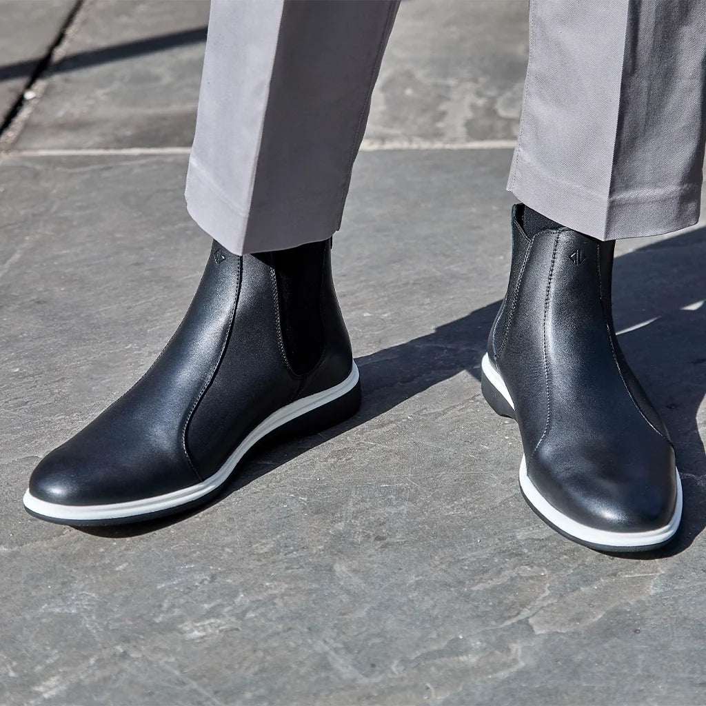 Black leather Chelsea boots with white soles worn by a person on a stone pavement.