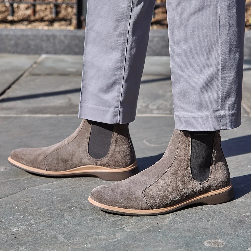 Gray suede Chelsea boots worn with light gray pants on a stone pavement.