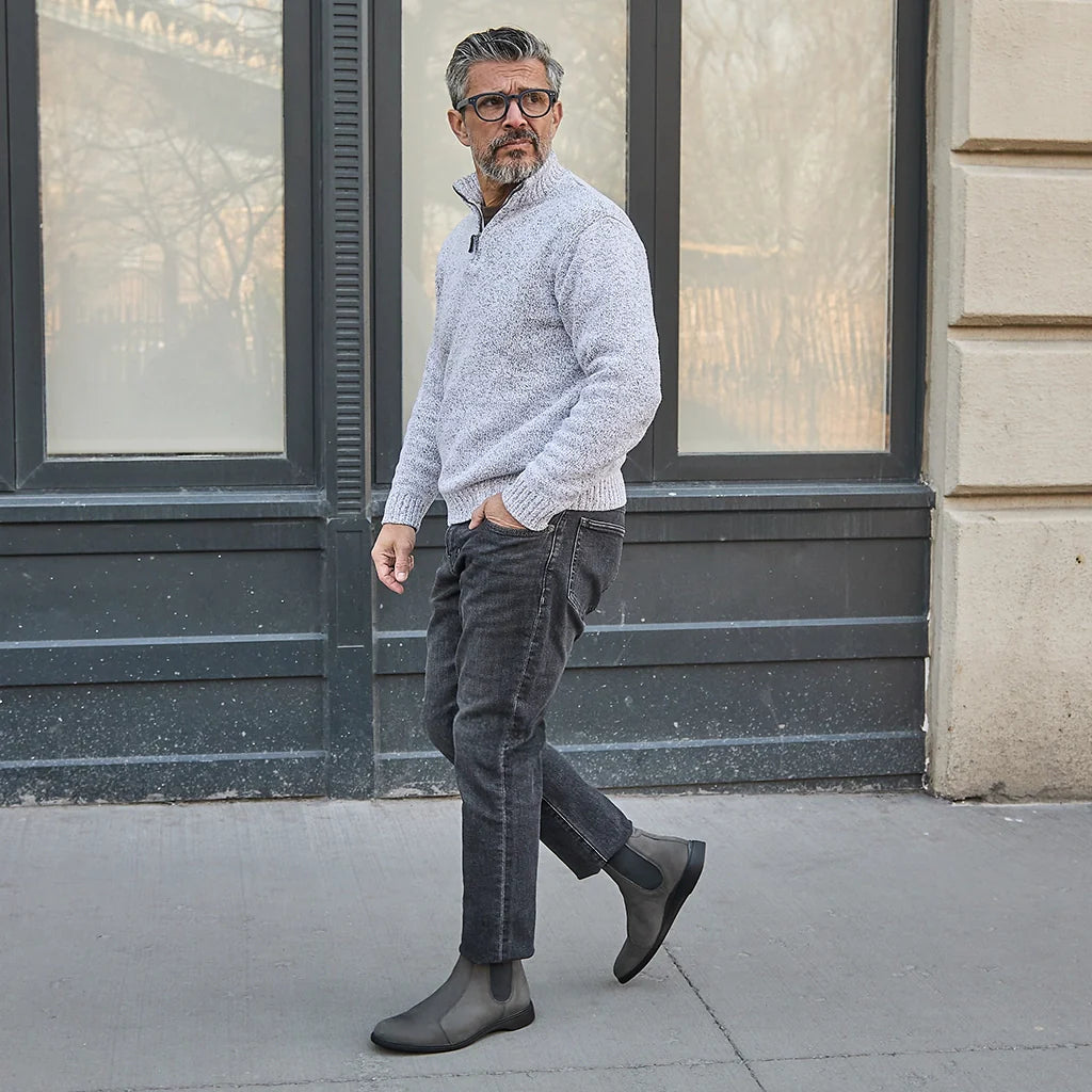 Man wearing a gray sweater, dark jeans, and black boots walking on a sidewalk.