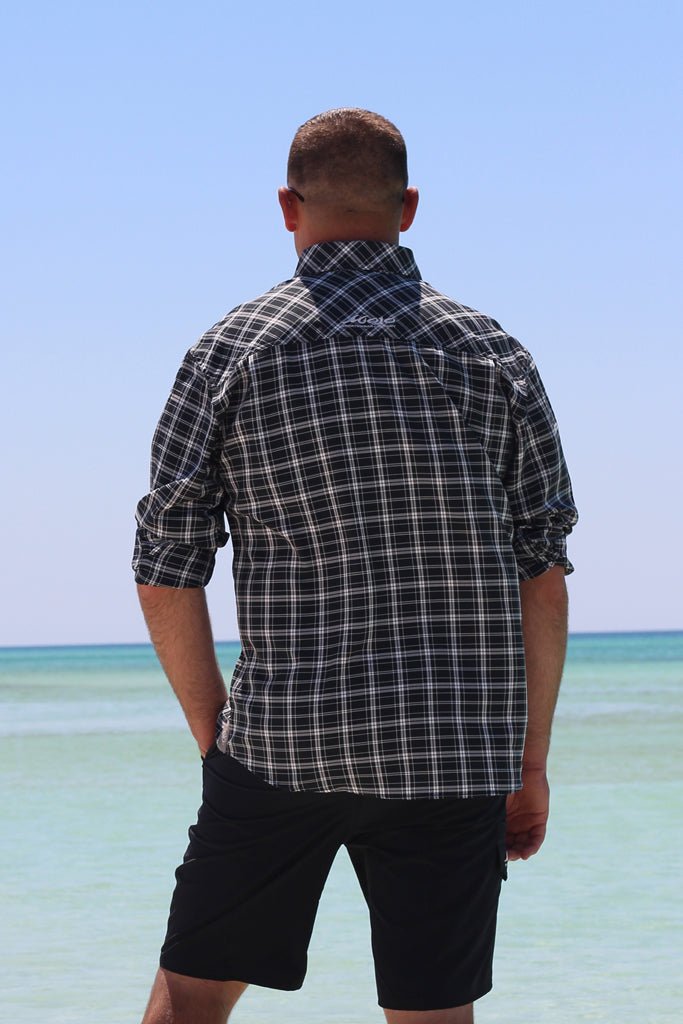 Man wearing a plaid shirt and black shorts standing on a beach with clear blue water and sky.