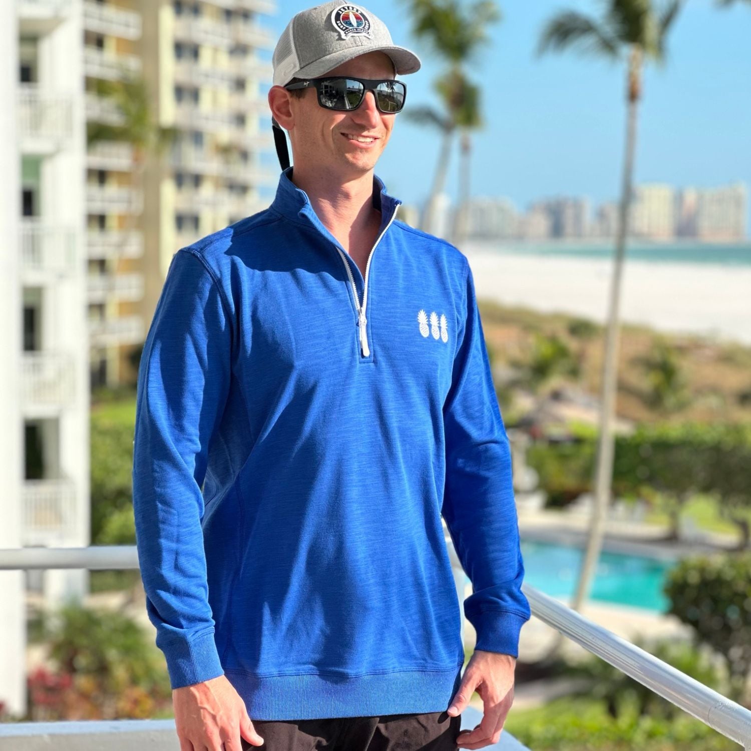Man wearing a blue pullover with a logo, sunglasses, and a cap in an outdoor setting with palm trees and buildings.