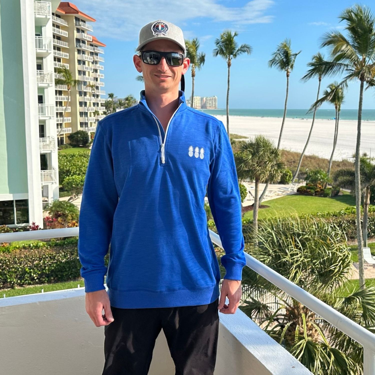 Man wearing a blue pullover with a logo, sunglasses, and a cap on a balcony with palm trees and beach in the background.