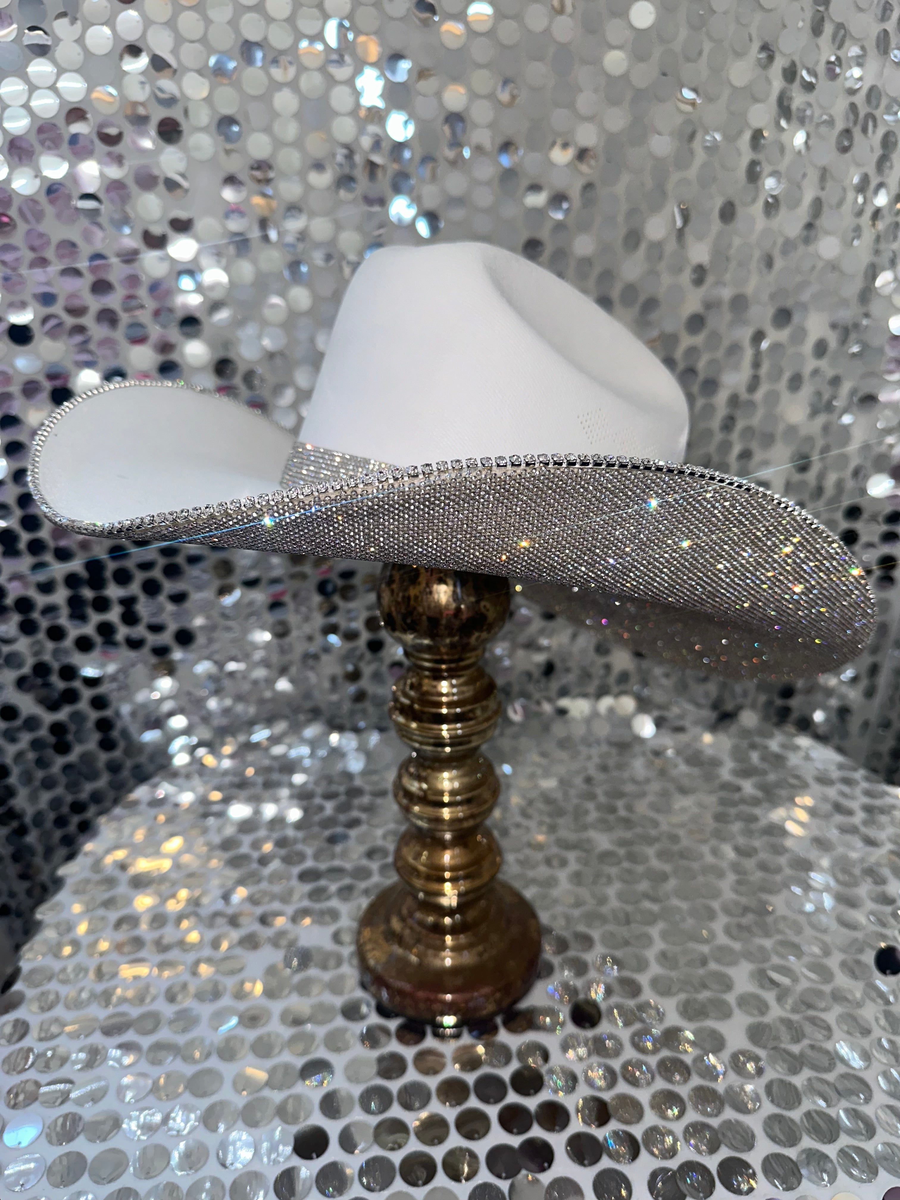 White cowboy hat with a silver brim on a gold stand against a silver sequin background