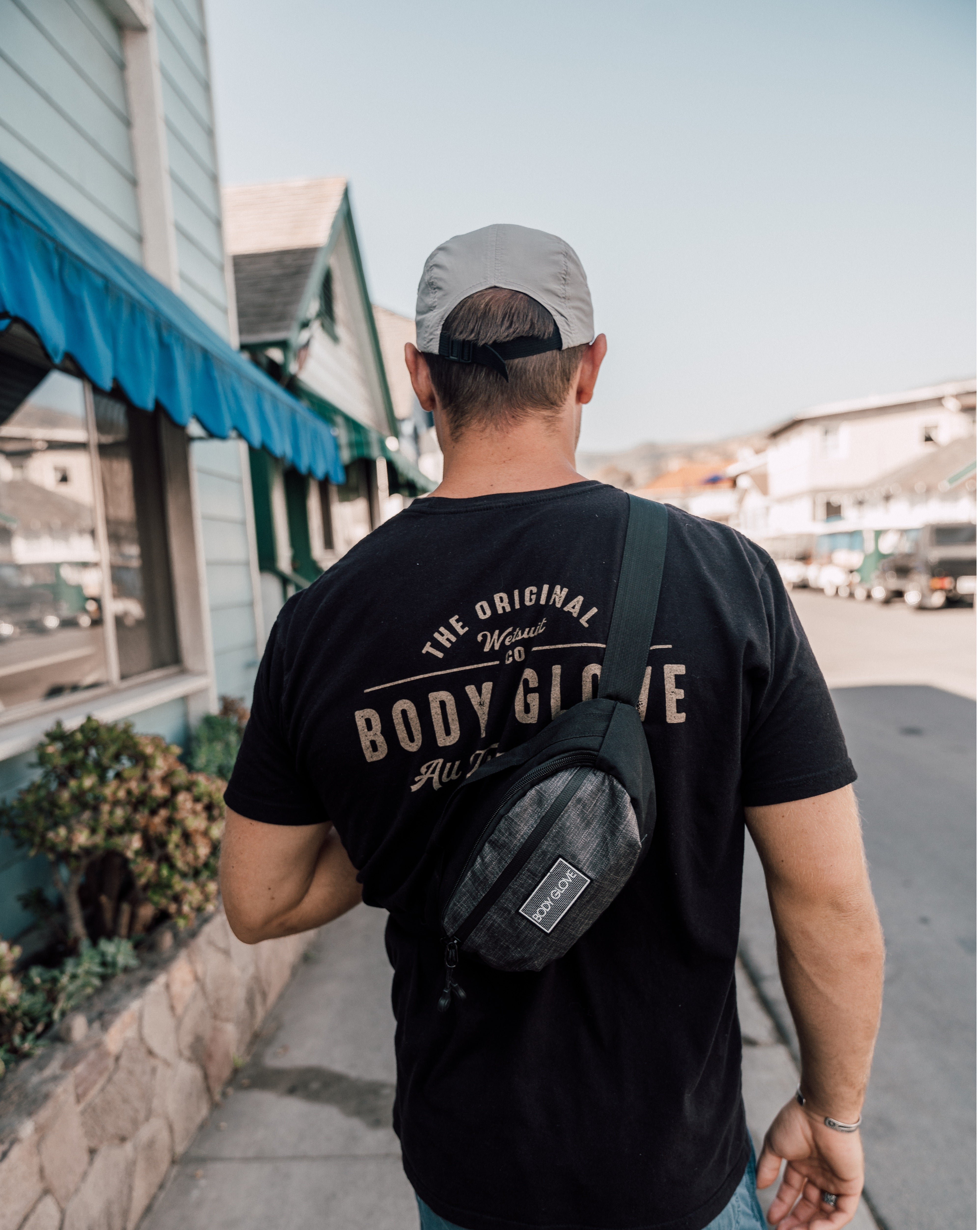 Man wearing a black 'Body Glove' t-shirt and carrying a black bag on a sidewalk.