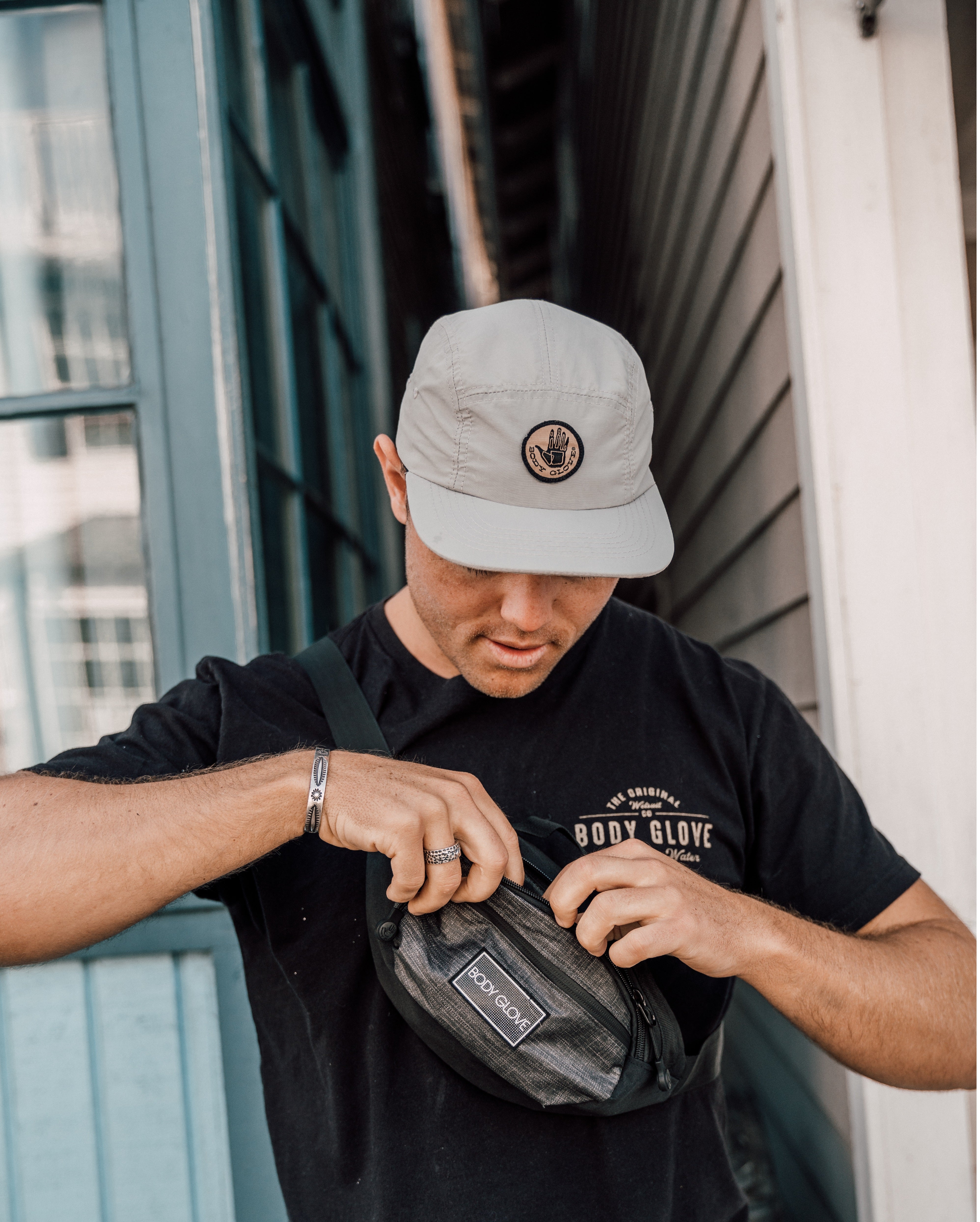 Person wearing a cap and black t-shirt with a logo, holding a black bag.