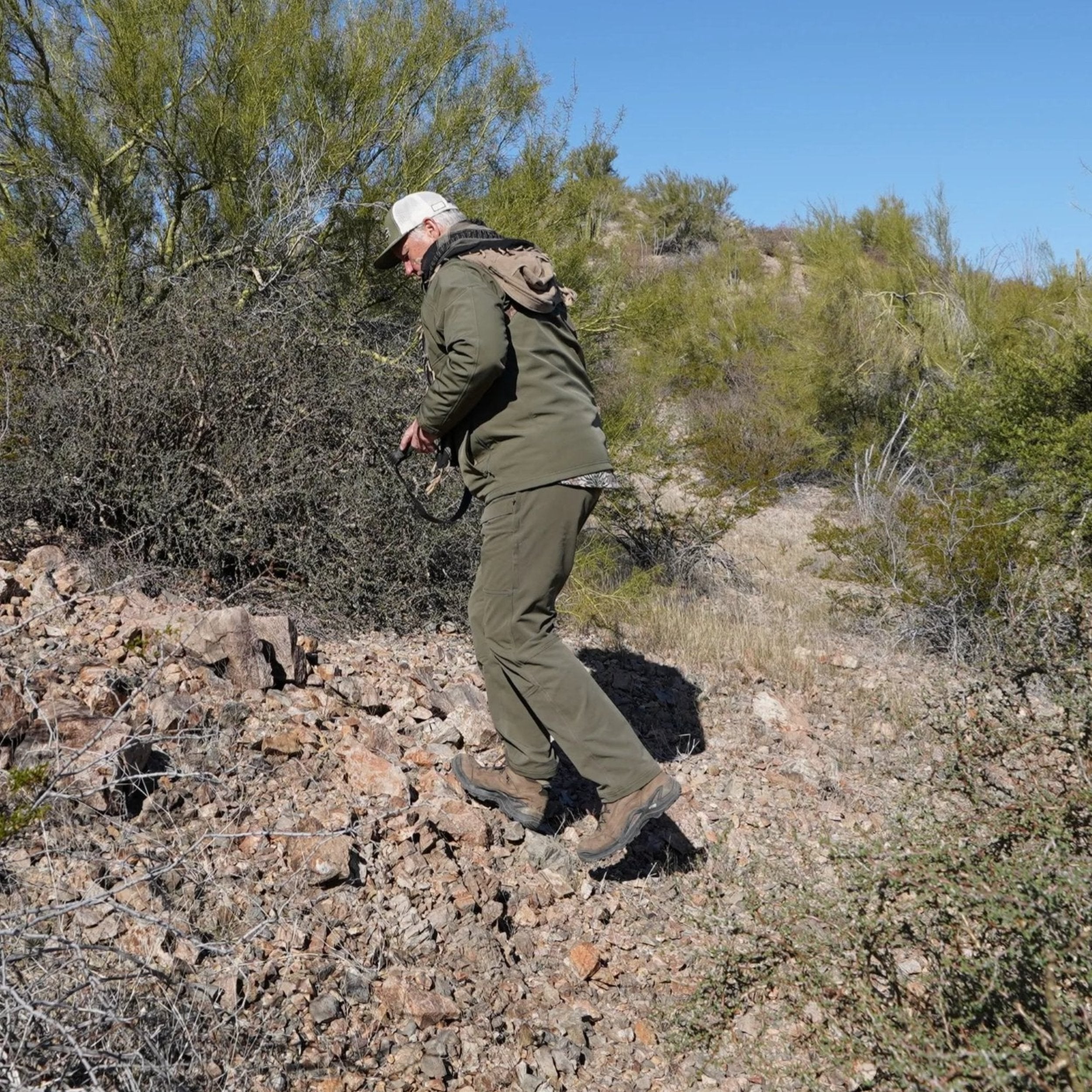 Person in green hunting gear walking on a rocky trail in a desert landscape