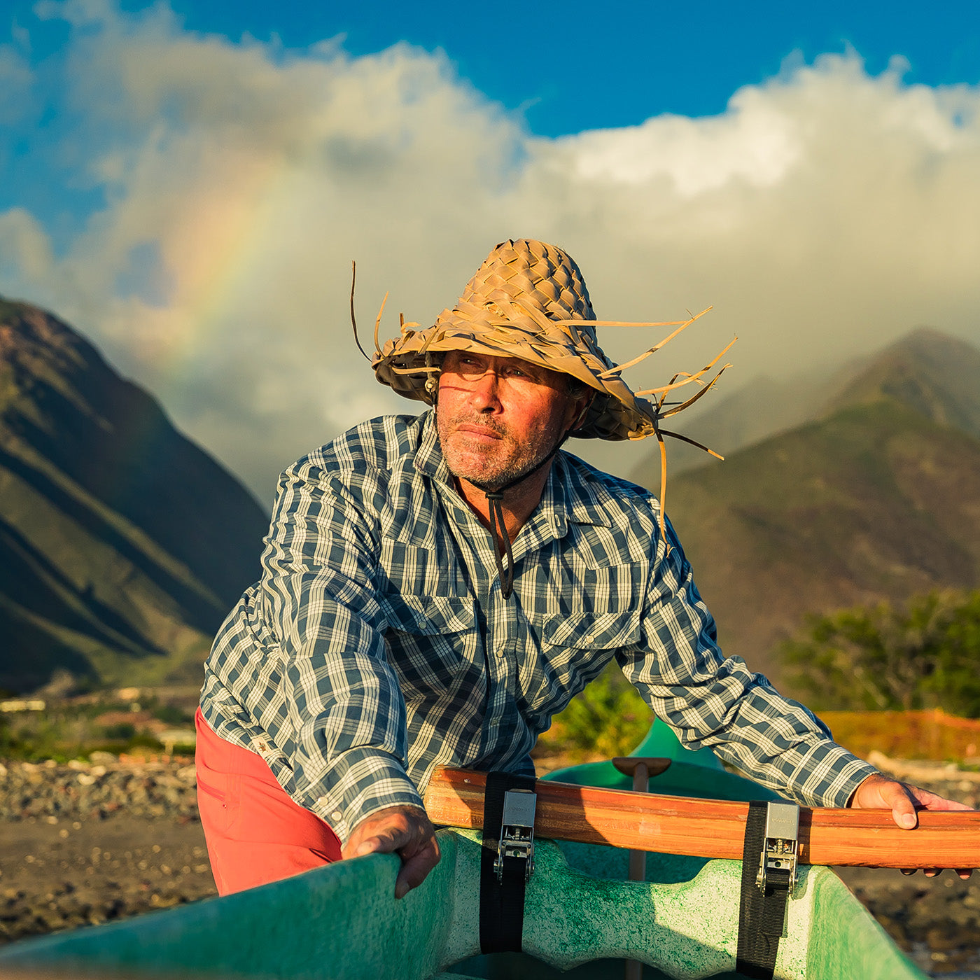 Man in a checkered shirt and straw hat sitting in a kayak with mountains and a rainbow in the background.