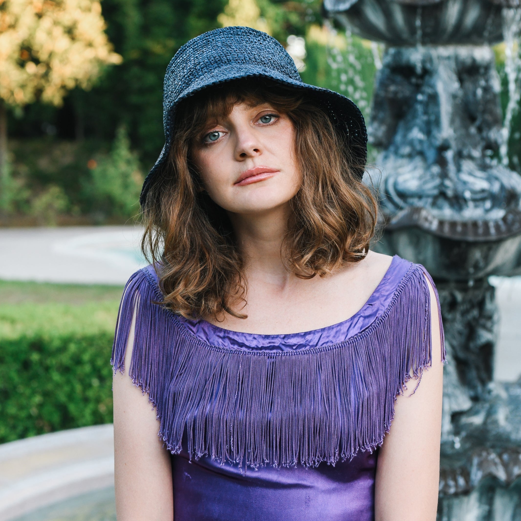 Woman wearing a blue hat and purple dress with fringe in an outdoor setting