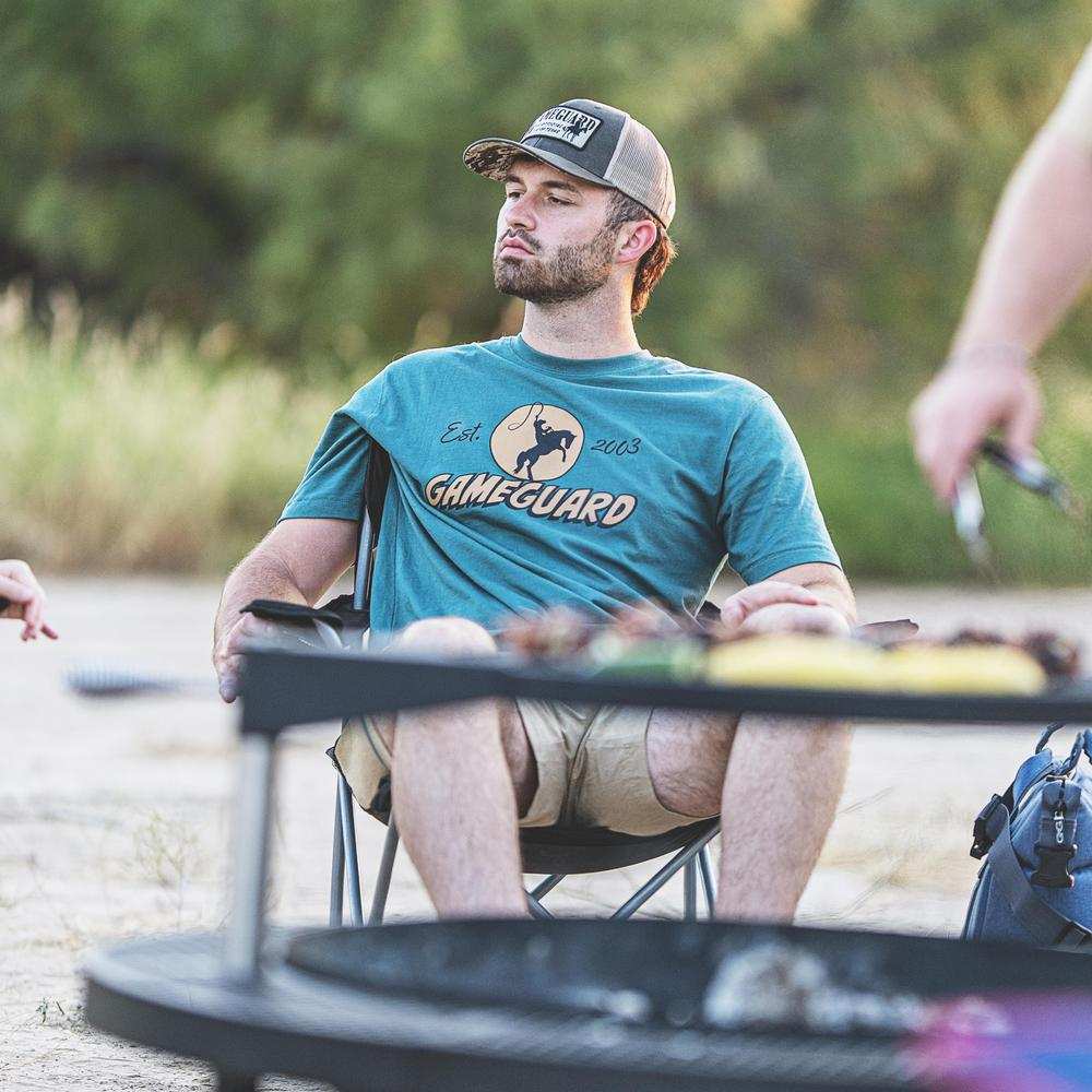 Man sitting outdoors by a campfire wearing a 'GameGuard' t-shirt.