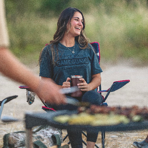 Woman sitting outdoors by a campfire, holding a mug with food on a grill in front of her.