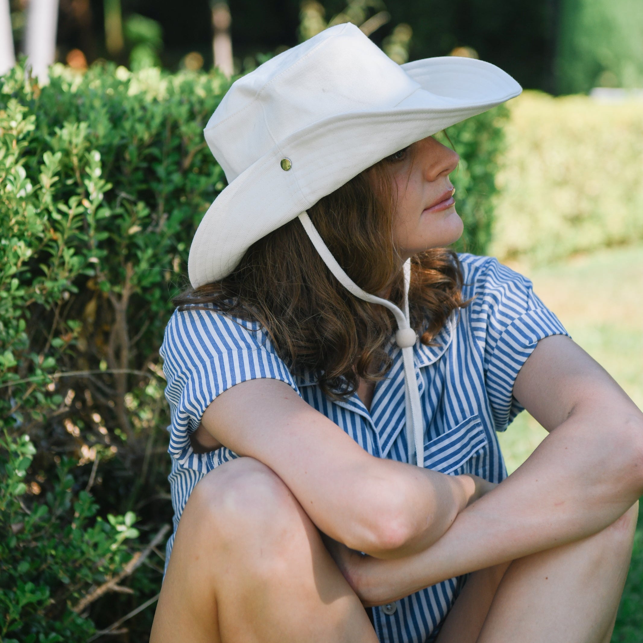 Woman wearing a white wide-brimmed hat sitting outdoors with greenery in the background