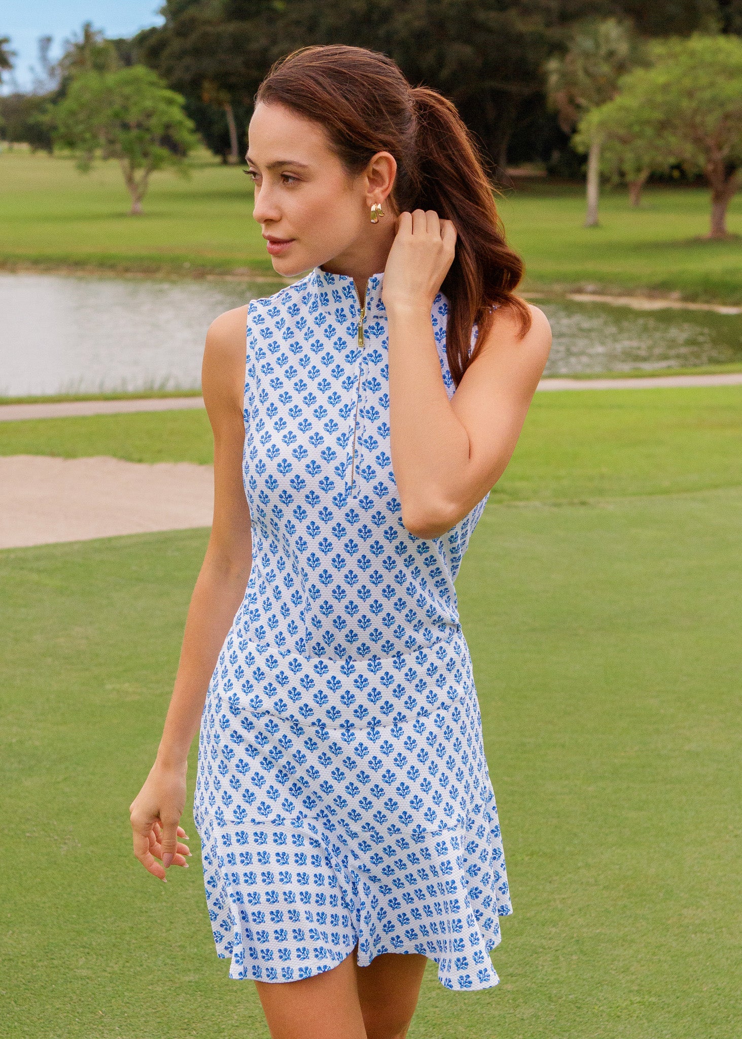 Woman in a blue patterned dress standing on a golf course with a pond and trees in the background.