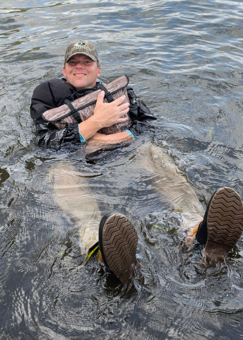 Person wading in water holding a large fish, wearing a cap and rain boots.