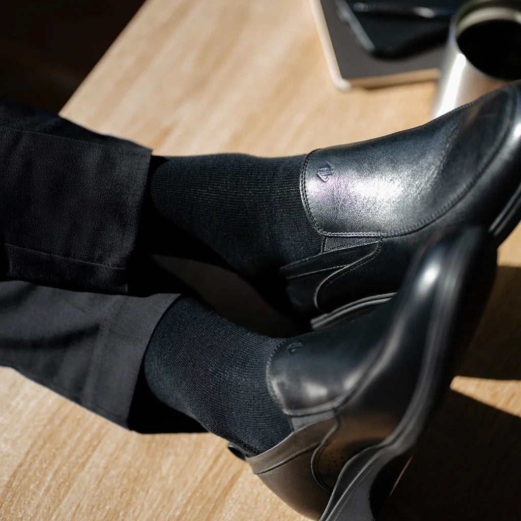 Person wearing black dress shoes and socks on a wooden floor.