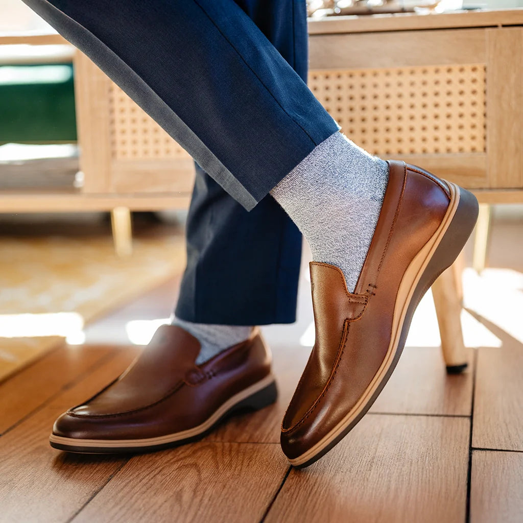 Brown leather shoes worn with blue pants and light gray socks on a wooden floor.