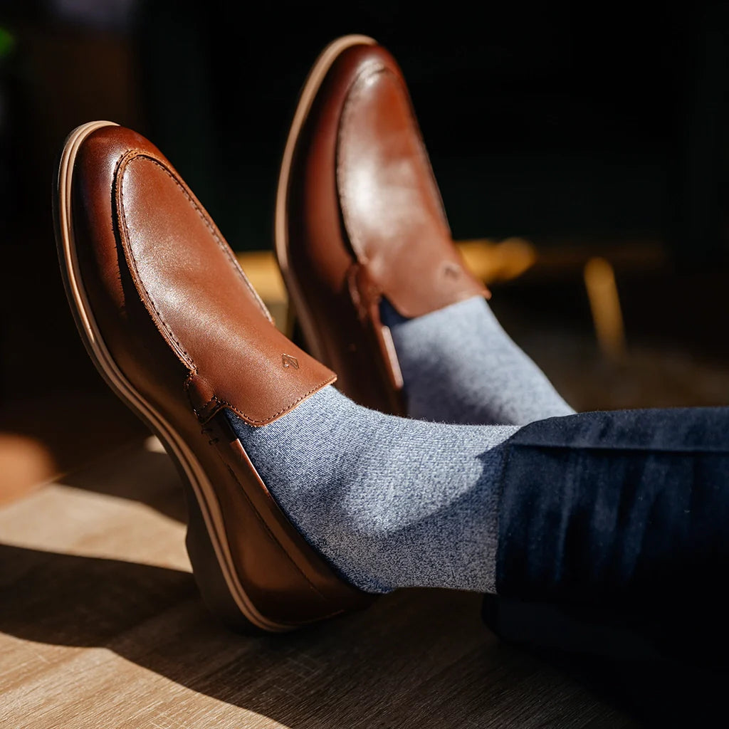 Brown leather loafers worn with blue socks and jeans on a wooden floor.